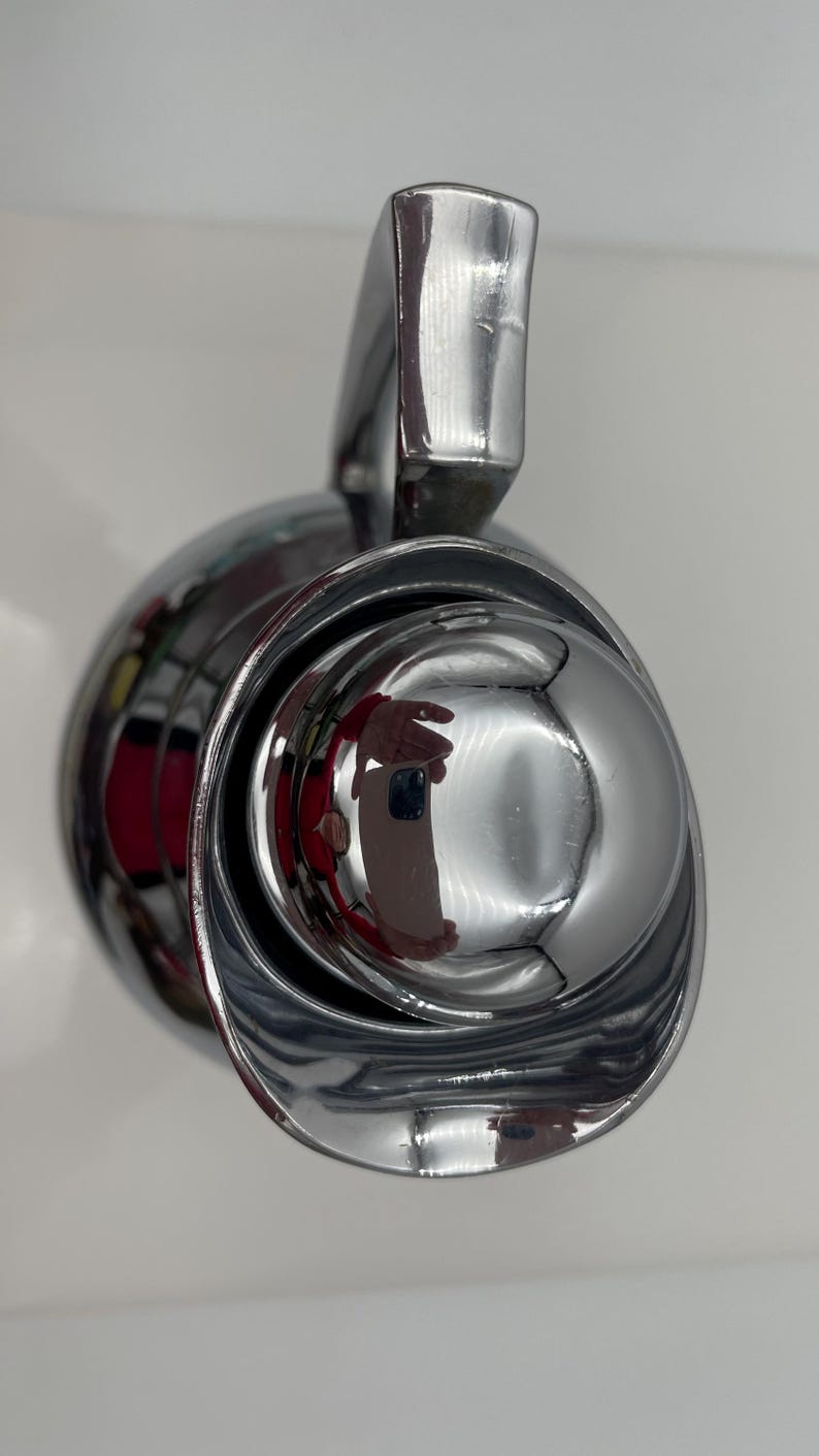 May include: A close-up of a shiny, chrome-plated object with a rounded, bowl-like shape and a curved handle. The surface reflects the surrounding environment, creating a distorted mirror effect. The object appears to be a faucet or similar fixture.