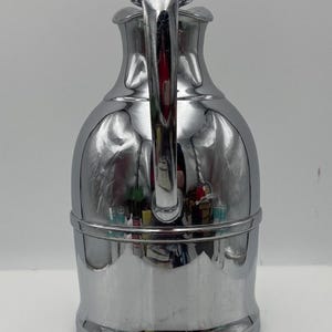 May include: A polished chrome jug with a curved handle and a rounded lid. The jug has a bulbous body and a narrow neck, with a horizontal band near the base. The surface reflects the surroundings, creating a mirror-like effect. A decorative item.