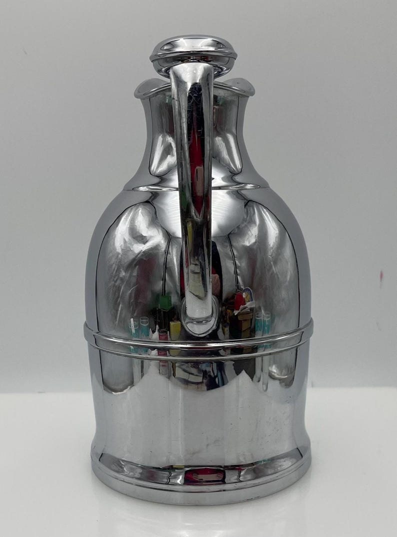 May include: A polished chrome jug with a curved handle and a rounded lid. The jug has a bulbous body and a narrow neck, with a horizontal band near the base. The surface reflects the surroundings, creating a mirror-like effect. A decorative item.