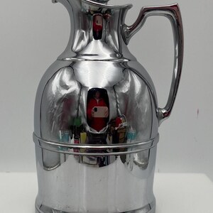 May include: A polished chrome vintage thermos flask with a lid and handle. The flask has a rounded body and a narrow neck, with a reflective surface. The handle is curved and attached to the side. The thermos is a classic design.