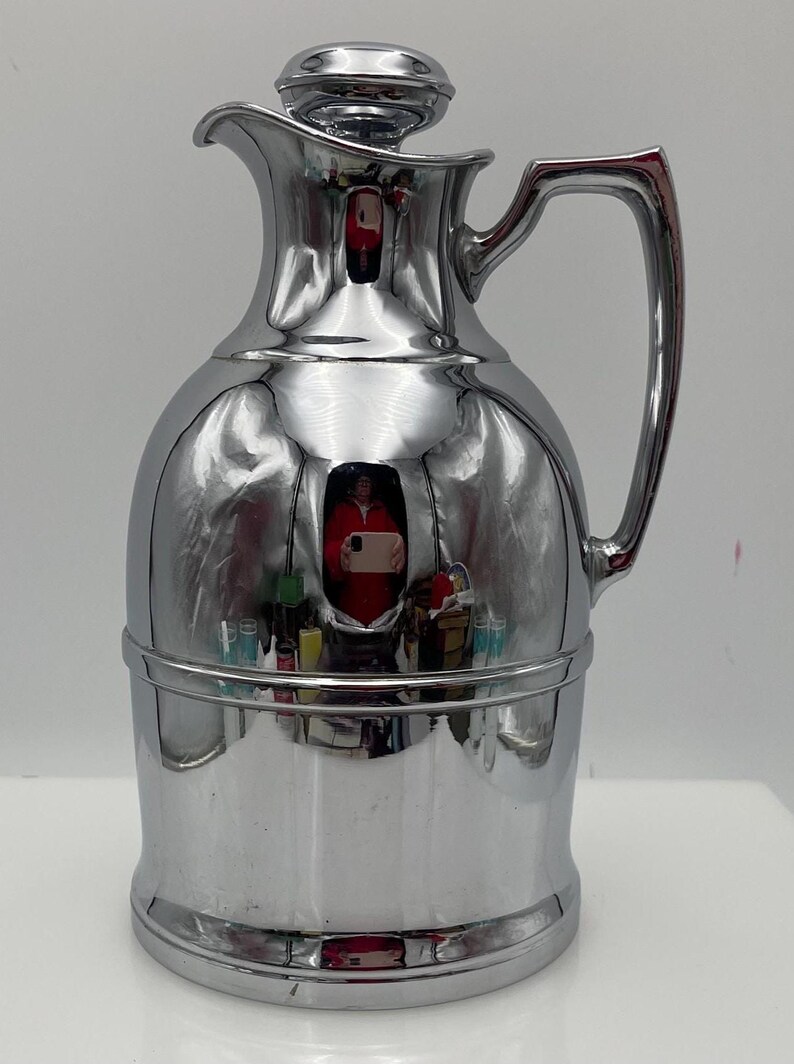 May include: A polished chrome vintage thermos flask with a lid and handle. The flask has a rounded body and a narrow neck, with a reflective surface. The handle is curved and attached to the side. The thermos is a classic design.