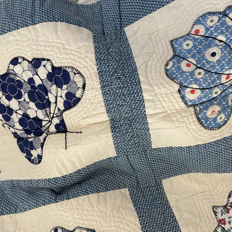 Peut inclure: Un textile matelass&eacute; pr&eacute;sentant un fond &agrave; motifs bleus et blancs avec des carr&eacute;s de couleur cr&egrave;me. Chaque carr&eacute; affiche une application en tissu en forme d'&eacute;ventail ou de coquillage, soulign&eacute;e par des coutures sombres et des lignes de matelassage.