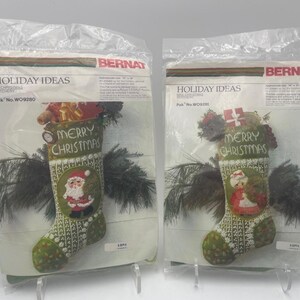 May include: Two packaged Christmas stocking kits. Each stocking is green with a white pattern, and features a Santa Claus or a girl in a red dress. The text "MERRY CHRISTMAS" is displayed on each stocking.
