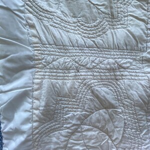 Peut inclure: Un textile matelass&eacute; blanc pr&eacute;sentant des motifs cousus complexes, notamment des lignes courbes et des formes g&eacute;om&eacute;triques. Le tissu poss&egrave;de une bordure lisse sur le c&ocirc;t&eacute; gauche et une surface textur&eacute;e et rembourr&eacute;e avec des coutures visibles.