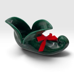 May include: A dark green ceramic shoe-shaped planter or decorative bowl with a glossy finish. The object features a curled toe and a wide, open top. A bright red fabric ribbon is tied in a bow around the front section of the shoe-shaped vessel.