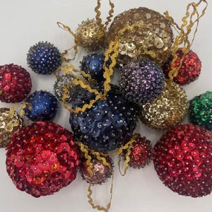 May include: A collection of decorative Christmas ornaments in various colors, including red, gold, blue, and green. The ornaments are round and covered in sequins, with gold ribbon hangers.