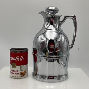 May include: A polished chrome vintage thermos flask with a lid and handle, reflecting the environment. Beside it is a can of Campbell's Cream of Mushroom Soup. The flask is a classic design, probably for keeping drinks hot or cold.