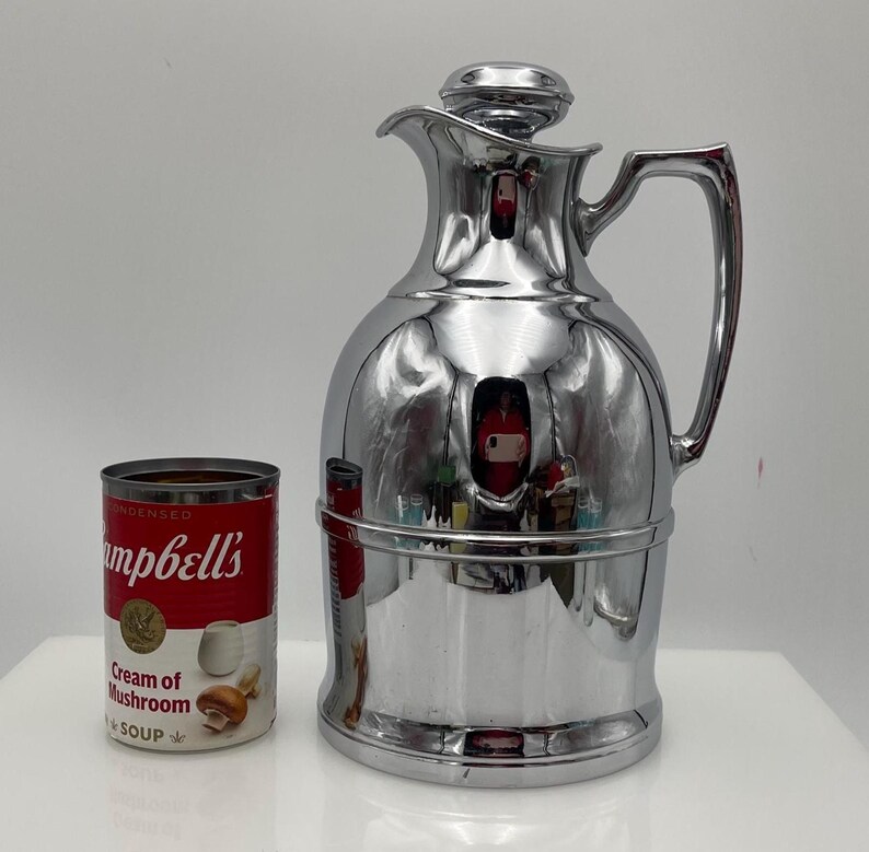 May include: A polished chrome vintage thermos flask with a lid and handle, reflecting the environment. Beside it is a can of Campbell's Cream of Mushroom Soup. The flask is a classic design, probably for keeping drinks hot or cold.