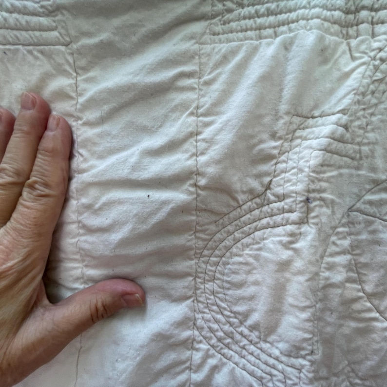 Peut inclure: Un textile en tissu blanc pr&eacute;sentant des lignes de couture parall&egrave;les complexes et des motifs de matelassage d&eacute;coratifs incurv&eacute;s. Le mat&eacute;riau a une texture douce et l&eacute;g&egrave;rement froiss&eacute;e avec des coutures visibles reliant les sections.