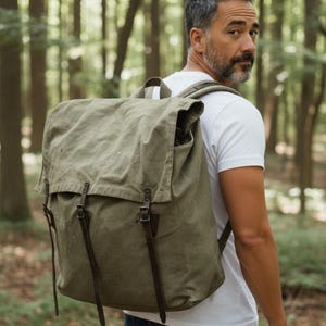 May include: An olive green canvas backpack with leather straps and buckles. The backpack has a large flap closure and is worn by a person in a white t-shirt and blue jeans. The background is a forest with tall trees.