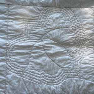 Peut inclure: Un tissu matelass&eacute; blanc pr&eacute;sentant un motif central cousu compos&eacute; de formes g&eacute;om&eacute;triques arrondies et concentriques. La surface offre un aspect doux et textur&eacute; avec des lignes de fil visibles formant un motif sym&eacute;trique r&eacute;p&eacute;titif sur le textile.
