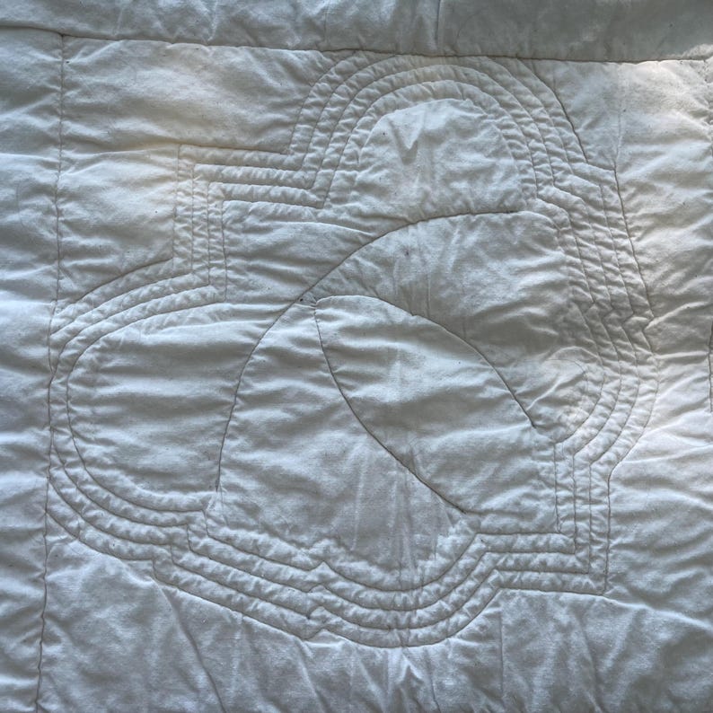 Peut inclure: Un tissu matelass&eacute; blanc pr&eacute;sentant un motif central cousu compos&eacute; de formes g&eacute;om&eacute;triques arrondies et concentriques. La surface offre un aspect doux et textur&eacute; avec des lignes de fil visibles formant un motif sym&eacute;trique r&eacute;p&eacute;titif sur le textile.