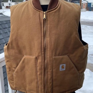 May include: A brown Carhartt vest with a brown collar and a full-length zipper. The vest has two large front pockets and a small Carhartt logo patch. The vest is displayed on a mannequin.
