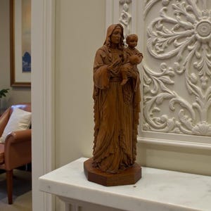 Könnte beinhalten: Eine Holzstatue einer bekleideten Figur, die ein Kind hält. Die Statue ist aus hellbraunem Holz geschnitzt und steht auf einem dunkelbraunen, sechseckigen Sockel. Im Hintergrund eine weiße Wand mit Zierleisten und ein Blick in einen Raum mit einem Stuhl.