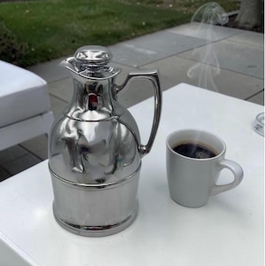 May include: A shiny, silver thermal carafe with a handle and a matching lid, next to a white mug filled with dark coffee. Steam rises from the coffee, suggesting it is hot. The items are on a white surface, possibly a table.