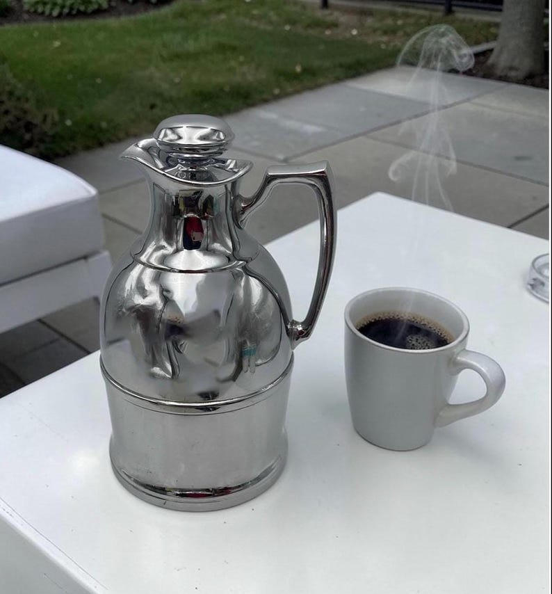 May include: A polished silver thermal flask with a handle and lid, alongside a white mug containing dark coffee. Steam rises from the coffee, indicating it is hot. The items are on a white table surface.