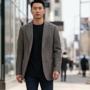 May include: A gray tweed blazer with a black crew neck shirt and dark blue jeans. The blazer has two front pockets and a notched lapel. The person is walking on a sidewalk in an urban setting.