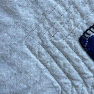 Peut inclure: Un gros plan sur un tissu matelass&eacute; blanc pr&eacute;sentant des rang&eacute;es parall&egrave;les de coutures droites formant un motif en angle. Une partie d'une pi&egrave;ce de tissu bleu fonc&eacute; &agrave; pois blancs est visible sur le bord droit, fix&eacute;e par des coutures sombres.