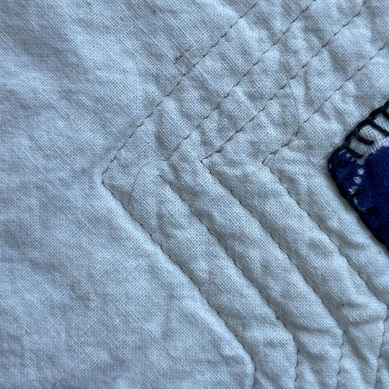Peut inclure: Un gros plan sur un tissu matelass&eacute; blanc pr&eacute;sentant des rang&eacute;es parall&egrave;les de coutures droites formant un motif en angle. Une partie d'une pi&egrave;ce de tissu bleu fonc&eacute; &agrave; pois blancs est visible sur le bord droit, fix&eacute;e par des coutures sombres.