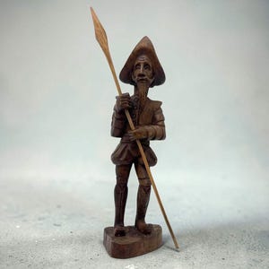 May include: A hand-carved wooden figurine of a man in historical attire, holding a spear. The figure has a long beard, a hat, and detailed clothing. The statue is brown and stands on a small wooden base. The spear is long and thin.