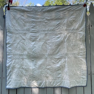 Peut inclure: Une courtepointe carr&eacute;e blanche compos&eacute;e d'une grille de neuf panneaux, chacun orn&eacute; de motifs de feuilles cousus complexes. La courtepointe est finie avec une fine bordure &agrave; motifs bleus et blancs et est maintenue par deux pinces rouges.