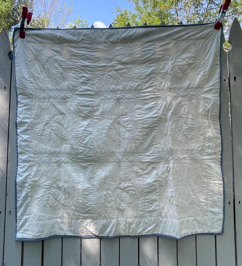 Peut inclure: Une courtepointe carr&eacute;e blanche compos&eacute;e d'une grille de neuf panneaux, chacun orn&eacute; de motifs de feuilles cousus complexes. La courtepointe est finie avec une fine bordure &agrave; motifs bleus et blancs et est maintenue par deux pinces rouges.