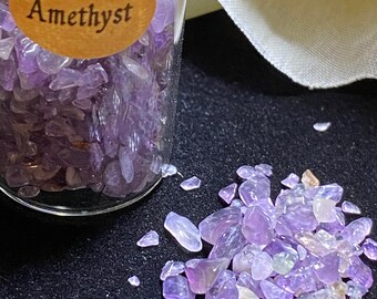 May include: A small glass jar filled with amethyst chips. The jar has a label that reads "Amethyst". Amethyst chips are scattered on a black surface.