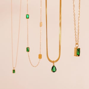Op de afbeelding: Vier gouden kettingen met groene edelsteen hangers. De kettingen hebben verschillende kettingstijlen, waaronder een delicate ketting, een dikkere ketting, een slangenketting en een ketting met een druppelhanger.