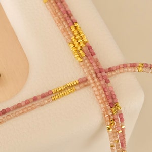 May include: Four strands of beaded necklaces in varying shades of pink and gold. The beads are small and square, with gold-colored accents. The necklaces are draped over a white surface.