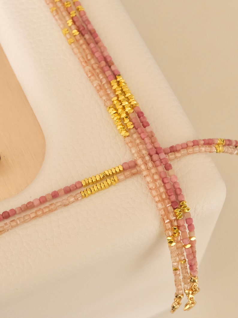 May include: Four strands of beaded necklaces in varying shades of pink and gold. The beads are small and square, with gold-colored accents. The necklaces are draped over a white surface.