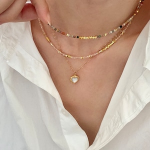 May include: A layered necklace set featuring three strands. The top strand has small, colorful beads, the middle strand has pink beads, and the bottom strand has a gold chain with a heart pendant. The heart pendant has a white center and gold trim.