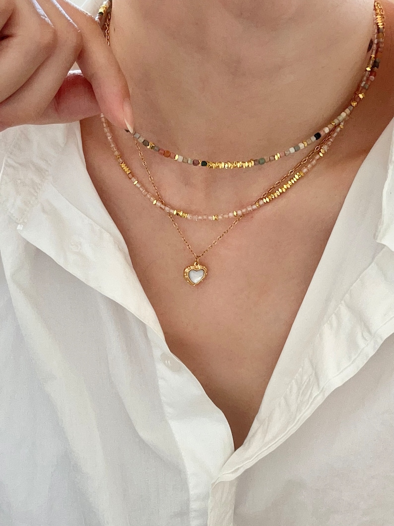 May include: A layered necklace set featuring three strands. The top strand has small, colorful beads, the middle strand has pink beads, and the bottom strand has a gold chain with a heart pendant. The heart pendant has a white center and gold trim.