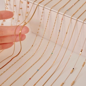 K&ouml;nnte beinhalten: Eine Sammlung von 15 goldenen Ketten in verschiedenen Stilen und L&auml;ngen, die auf einer wei&szlig;en Oberfl&auml;che pr&auml;sentiert werden. Einige Ketten haben kleine Perlen oder Anh&auml;nger.