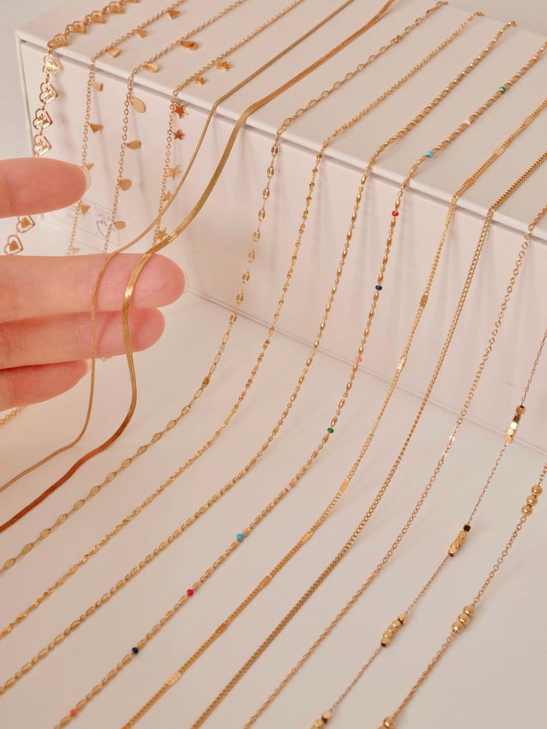 K&ouml;nnte beinhalten: Eine Sammlung von 15 goldenen Ketten in verschiedenen Stilen und L&auml;ngen, die auf einer wei&szlig;en Oberfl&auml;che pr&auml;sentiert werden. Einige Ketten haben kleine Perlen oder Anh&auml;nger.