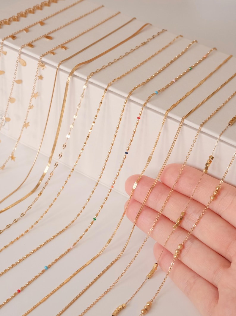 K&ouml;nnte beinhalten: Eine Sammlung von goldenen Kettenhalsketten mit verschiedenen Stilen und Verzierungen, darunter Perlen, Quadrate und kleine farbige Steine.
