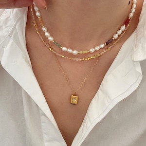 May include: A layered necklace set featuring three strands. The top strand has white pearls and colorful beads. The middle strand has pink and gold beads. The bottom strand has a gold pendant with a star design.