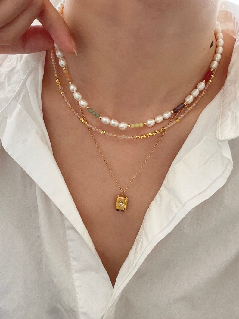 May include: A layered necklace set featuring three strands. The top strand has white pearls and colorful beads. The middle strand has pink and gold beads. The bottom strand has a gold pendant with a star design.