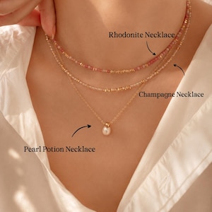 May include: Three necklaces are displayed. The top necklace is a Rhodonite Necklace with pink beads. The middle necklace is a Champagne Necklace with gold beads. The bottom necklace is a Pearl Potion Necklace with a single pearl pendant. All necklaces have gold chains.