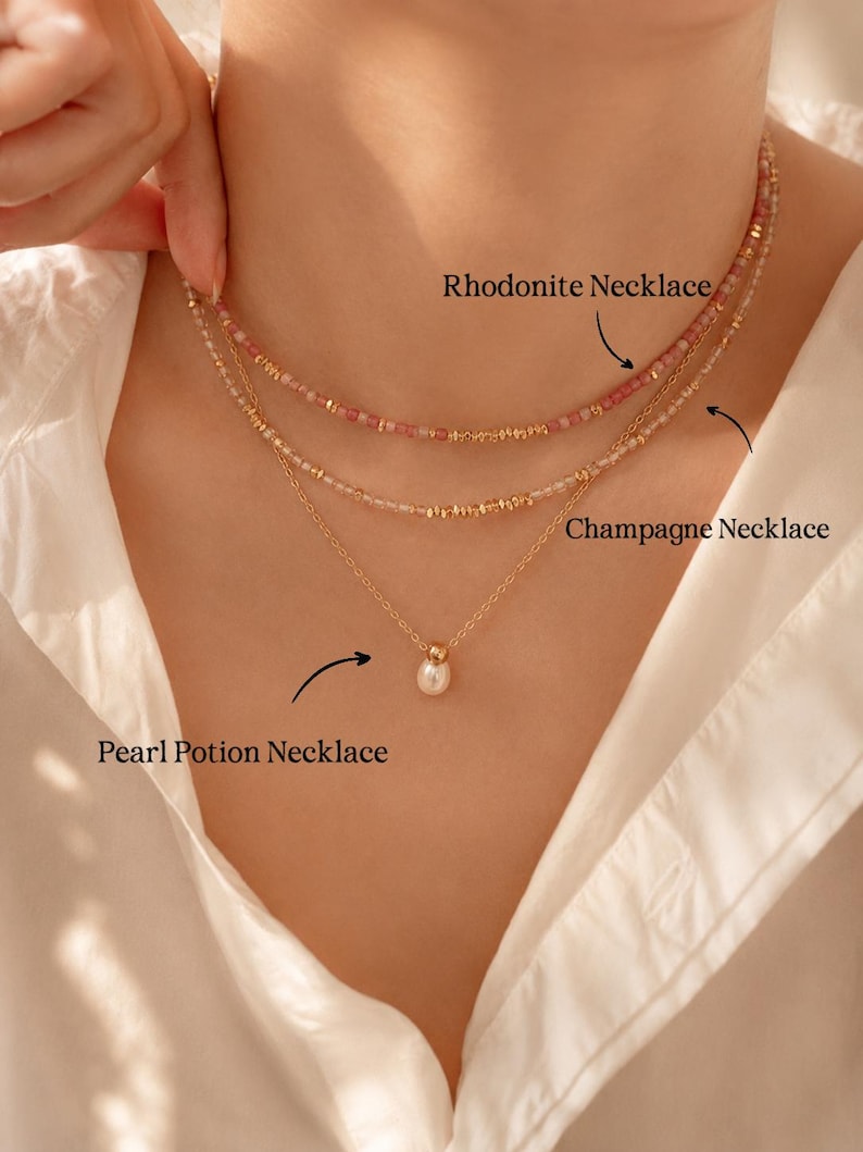 May include: Three necklaces are displayed. The top necklace is a Rhodonite Necklace with pink beads. The middle necklace is a Champagne Necklace with gold beads. The bottom necklace is a Pearl Potion Necklace with a single pearl pendant. All necklaces have gold chains.