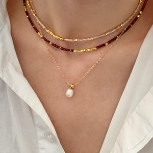May include: A layered necklace set featuring three strands. The top two strands have small beads in burgundy and pale pink, interspersed with gold-colored accents. The bottom strand is a delicate gold chain with a single pearl pendant.