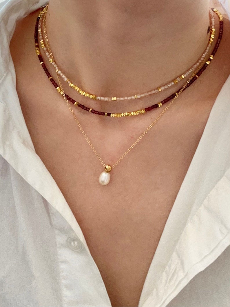 May include: A layered necklace set featuring three strands. The top two strands have small beads in burgundy and pale pink, interspersed with gold-colored accents. The bottom strand is a delicate gold chain with a single pearl pendant.