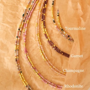 May include: Close-up of several delicate necklaces with small, colorful beads and gold accents. The necklaces are labeled with the names of the gemstones: Tourmaline, Garnet, Champagne, Rhodonite, and Agate. The necklaces are displayed on a brown paper background.