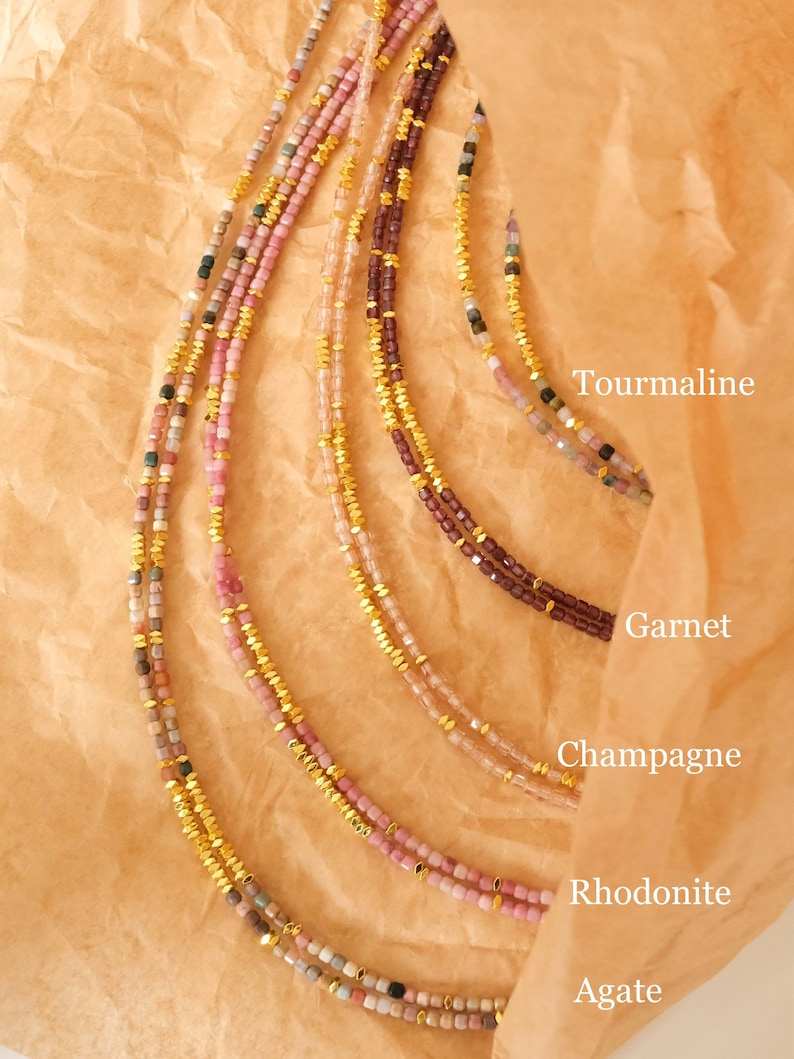 May include: Close-up of several delicate necklaces with small, colorful beads and gold accents. The necklaces are labeled with the names of the gemstones: Tourmaline, Garnet, Champagne, Rhodonite, and Agate. The necklaces are displayed on a brown paper background.