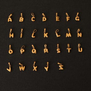 K&ouml;nnte beinhalten: Ein Satz von 26 goldfarbenen Alphabet-Anh&auml;ngern. Jeder Buchstabe ist ein separater Anh&auml;nger und wird auf einem schwarzen Hintergrund angezeigt.