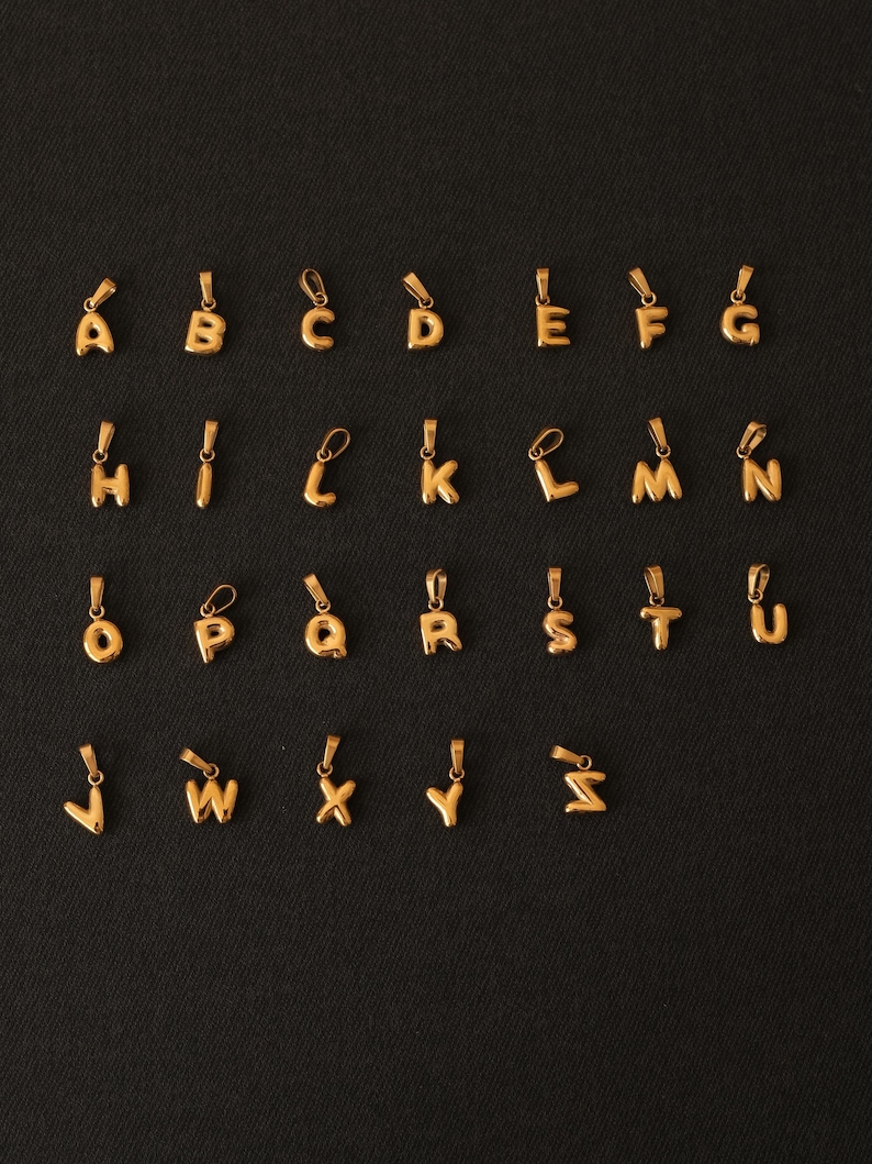 K&ouml;nnte beinhalten: Ein Satz von 26 goldfarbenen Alphabet-Anh&auml;ngern. Jeder Buchstabe ist ein separater Anh&auml;nger und wird auf einem schwarzen Hintergrund angezeigt.