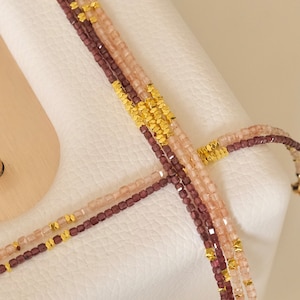May include: Close-up of multiple strands of beaded necklaces. The necklaces feature small, square beads in shades of mauve and light pink, interspersed with gold-colored metal accents. The necklaces are displayed on a white surface.