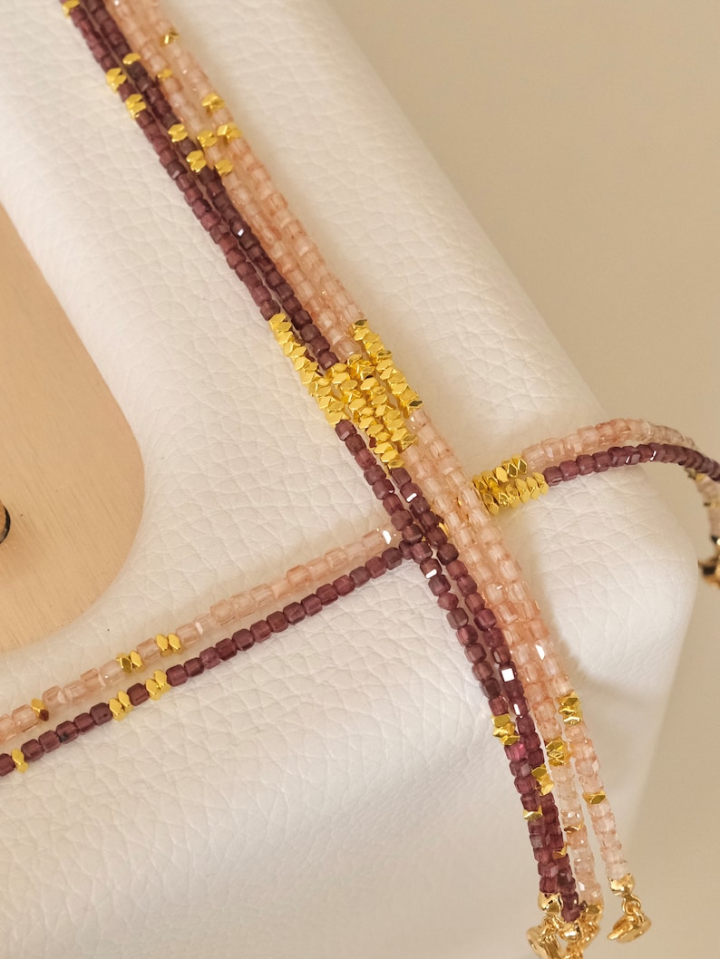 May include: Close-up of multiple strands of beaded necklaces. The necklaces feature small, square beads in shades of mauve and light pink, interspersed with gold-colored metal accents. The necklaces are displayed on a white surface.