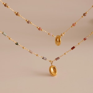 May include: Two gold necklaces with different pendant styles. The top necklace has a round pendant and the bottom necklace has an oval pendant. Both necklaces feature small, colourful beads.