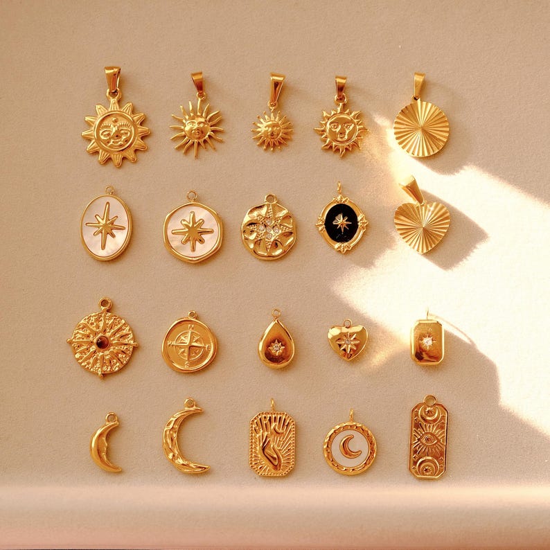 K&ouml;nnte beinhalten: Eine Sammlung von 19 goldfarbenen Anh&auml;ngern mit verschiedenen himmlischen und geometrischen Designs. Die Anh&auml;nger umfassen Sonnen, Sterne, Monde, Herzen und Kompassrosen.