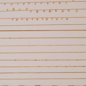 K&ouml;nnte beinhalten: Eine Sammlung von 12 goldenen Ketten mit verschiedenen Stilen und Designs. Die Ketten sind in Reihen auf einem wei&szlig;en Hintergrund angeordnet. Einige Ketten haben kleine Anh&auml;nger, wie Herzen, Sterne und Tropfen.